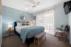 Ein Schlafzimmer mit einem Bett, Nachttischen, einer Bank, einem Sessel, einem Deckenventilator und einem an der Wand montierten Fernseher.