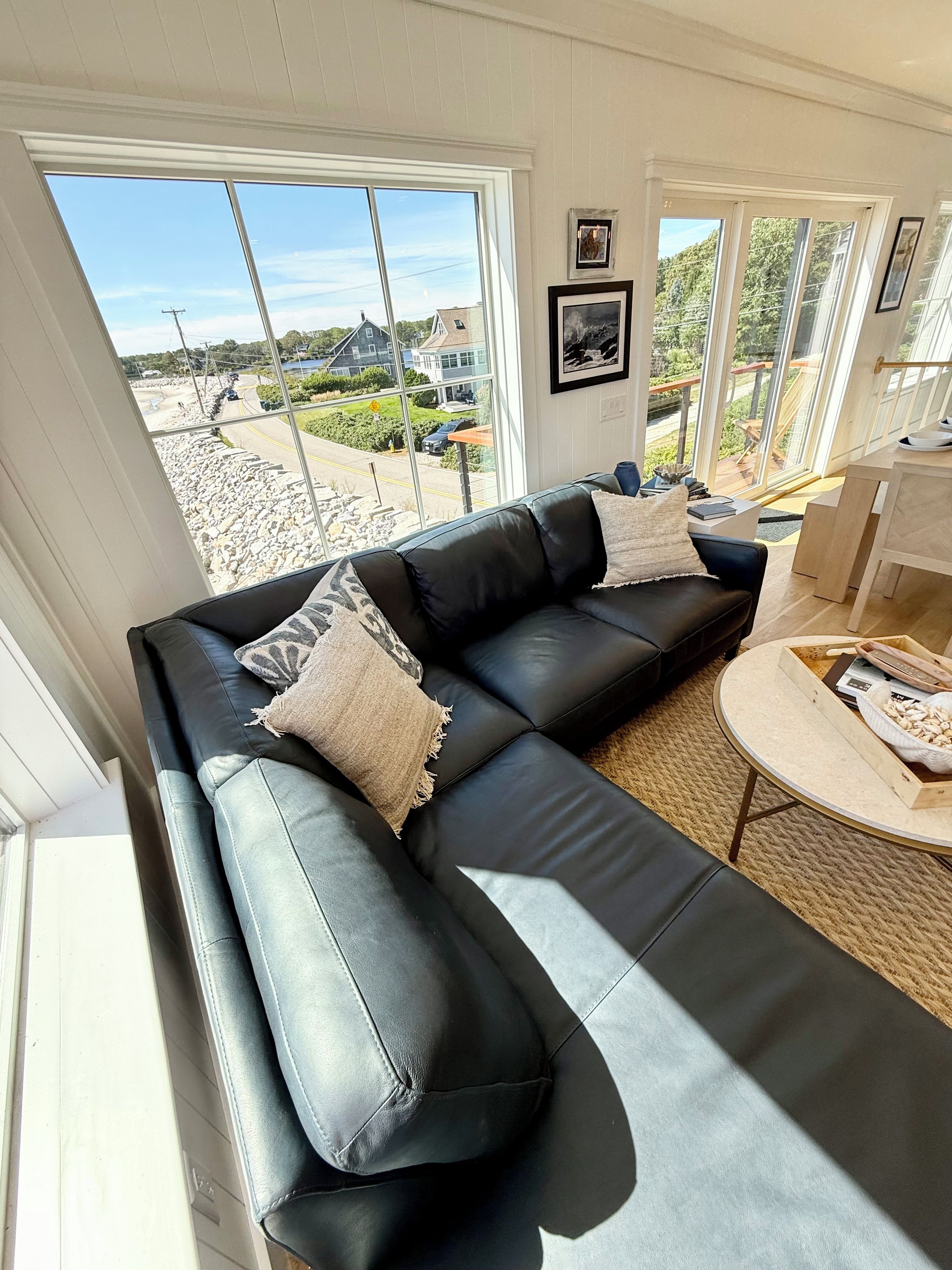 A modern living room with a black leather sofa, a round coffee table, and a large window offering a view of the outdoors.