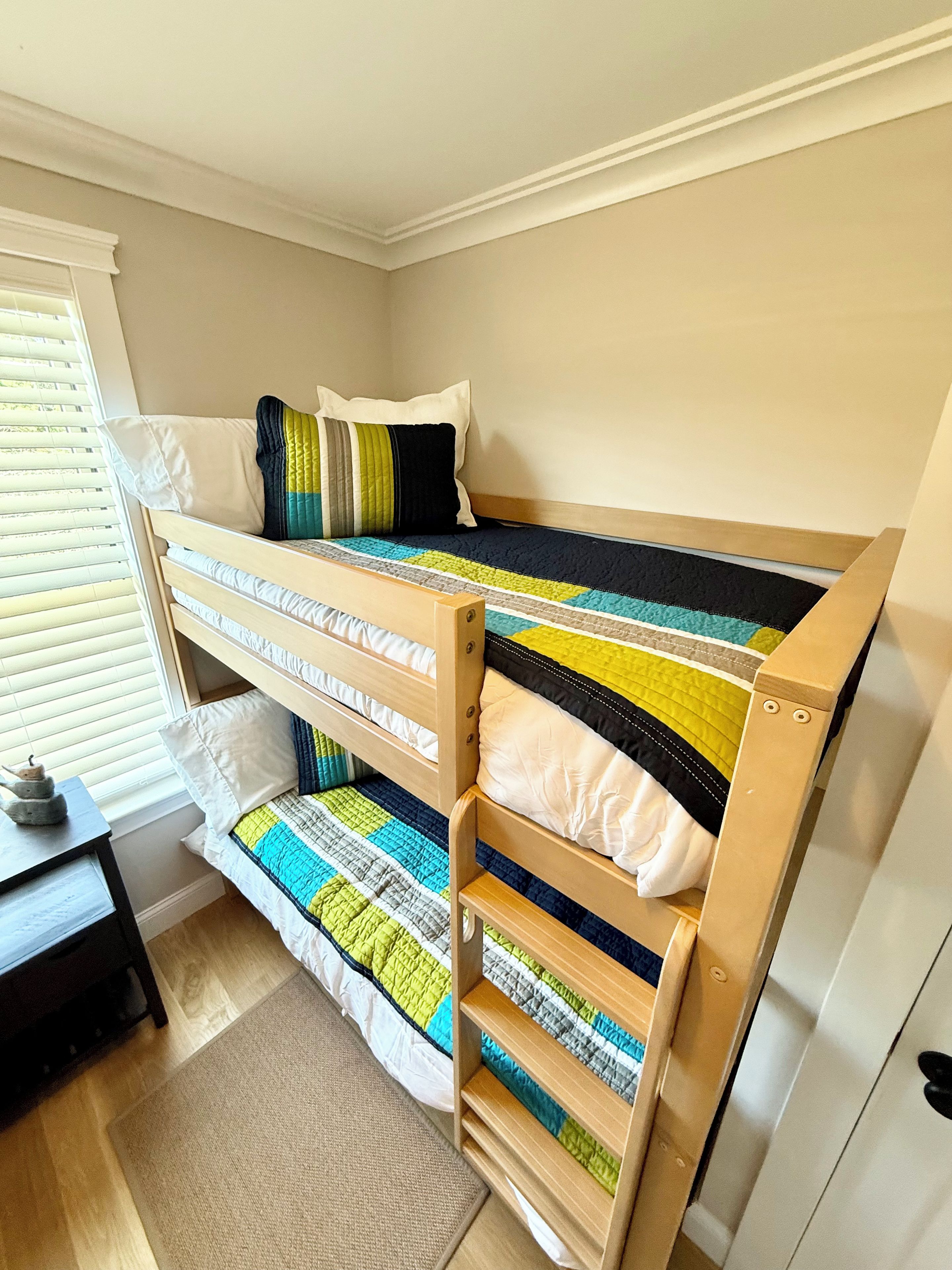 A bunk bed with a quilted bedspread, a wooden headboard, and a window with blinds.