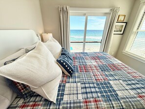 A bedroom with a bed, pillows, a view of the beach, and a window with blinds.