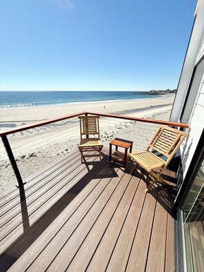 A wooden deck with two chairs and a small table, overlooking a beach with a clear sky.