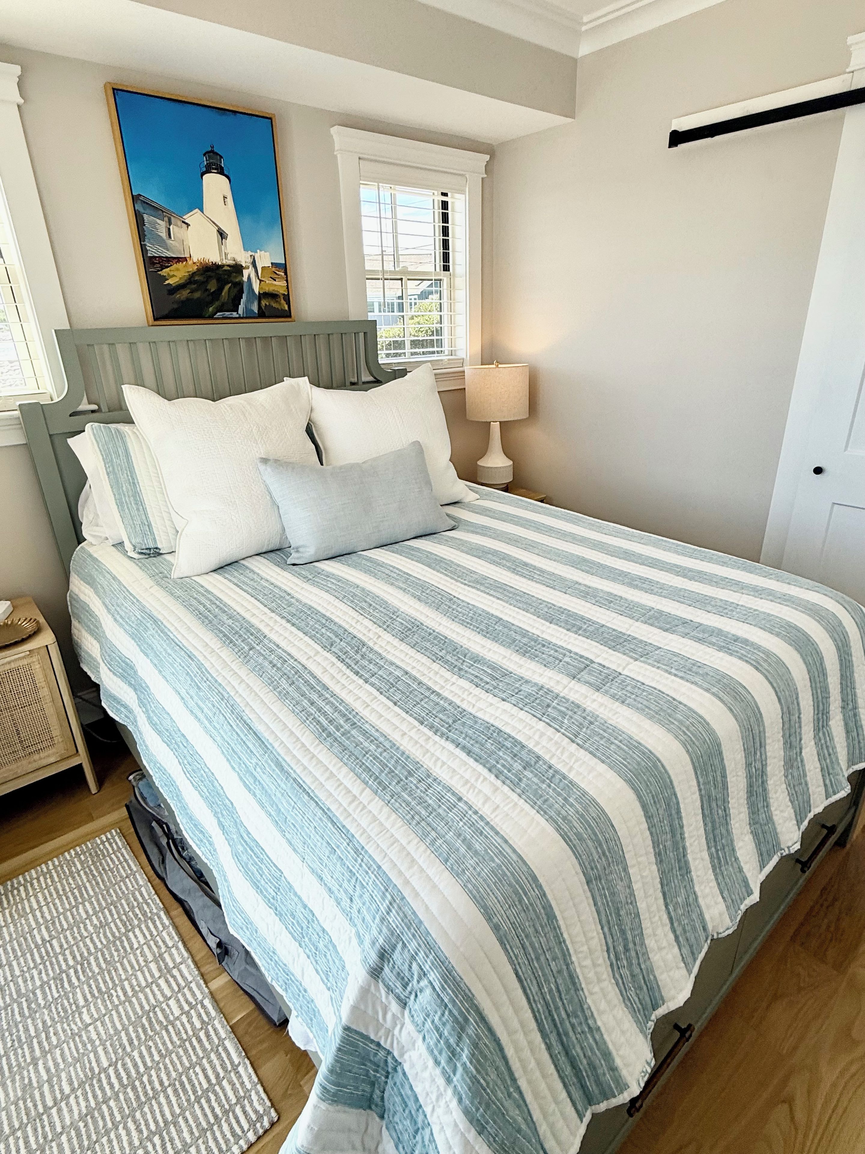 A bedroom with a bed, pillows, a nightstand, a lamp, a painting, and a window.