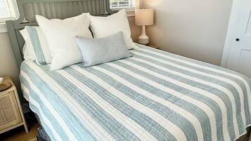 A bedroom with a bed, pillows, a nightstand, a lamp, a painting, and a window.