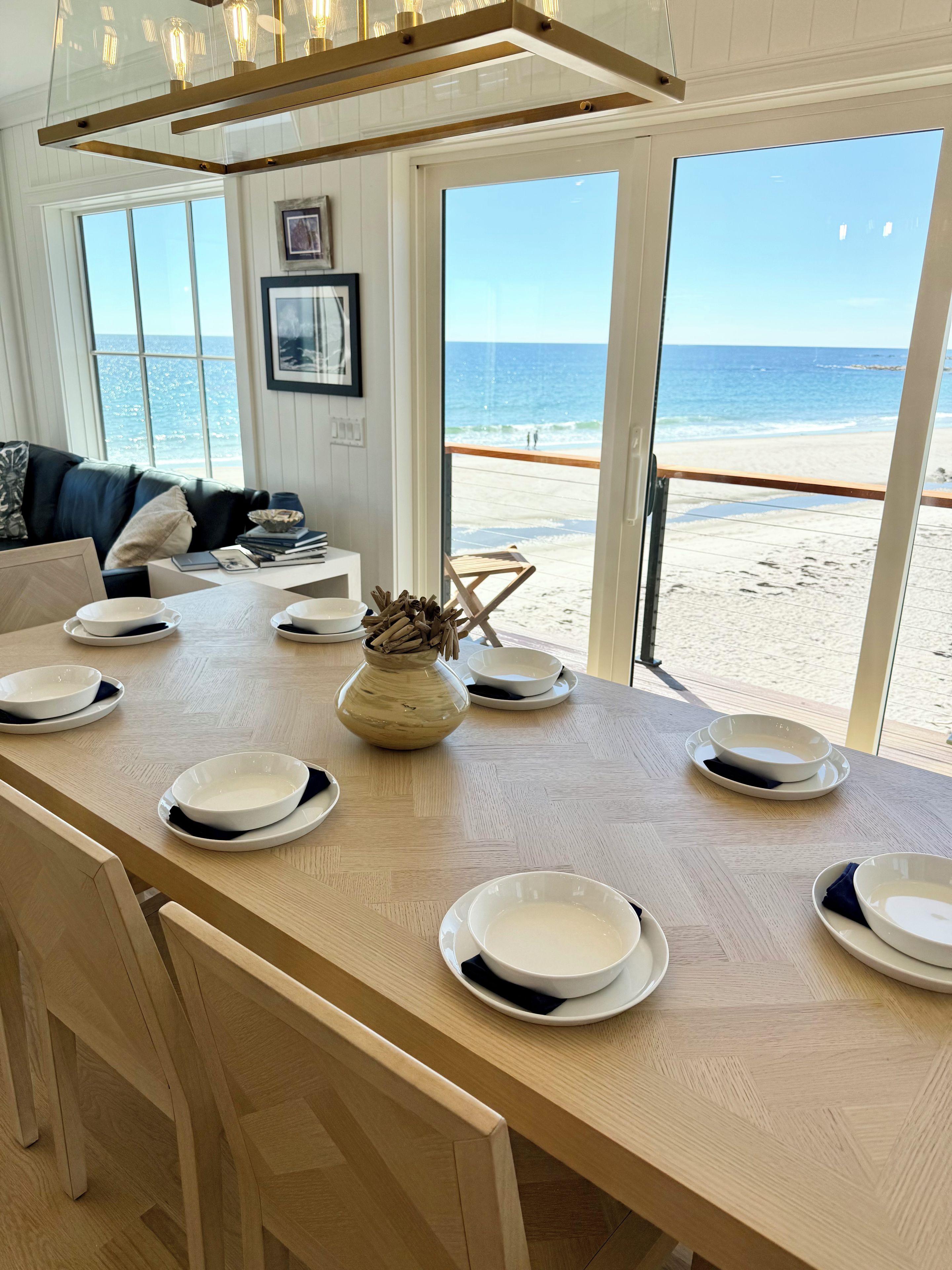 A dining area with a view of the beach, a wooden table set with plates, and a vase with dried flowers.