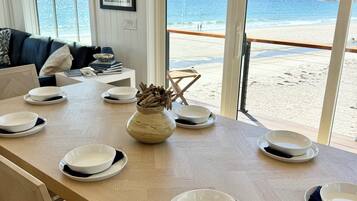 A dining area with a view of the beach, a wooden table set with plates, and a vase with dried flowers.