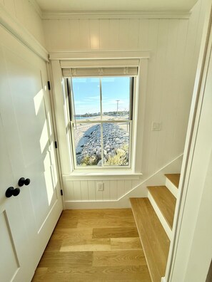 A room with a wooden floor, a window with a view of a rocky beach, and a white door with a handle.