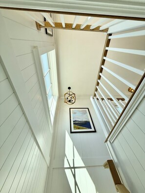 A narrow hallway with white walls and wooden beams, a framed picture on the wall, and a hanging light fixture.