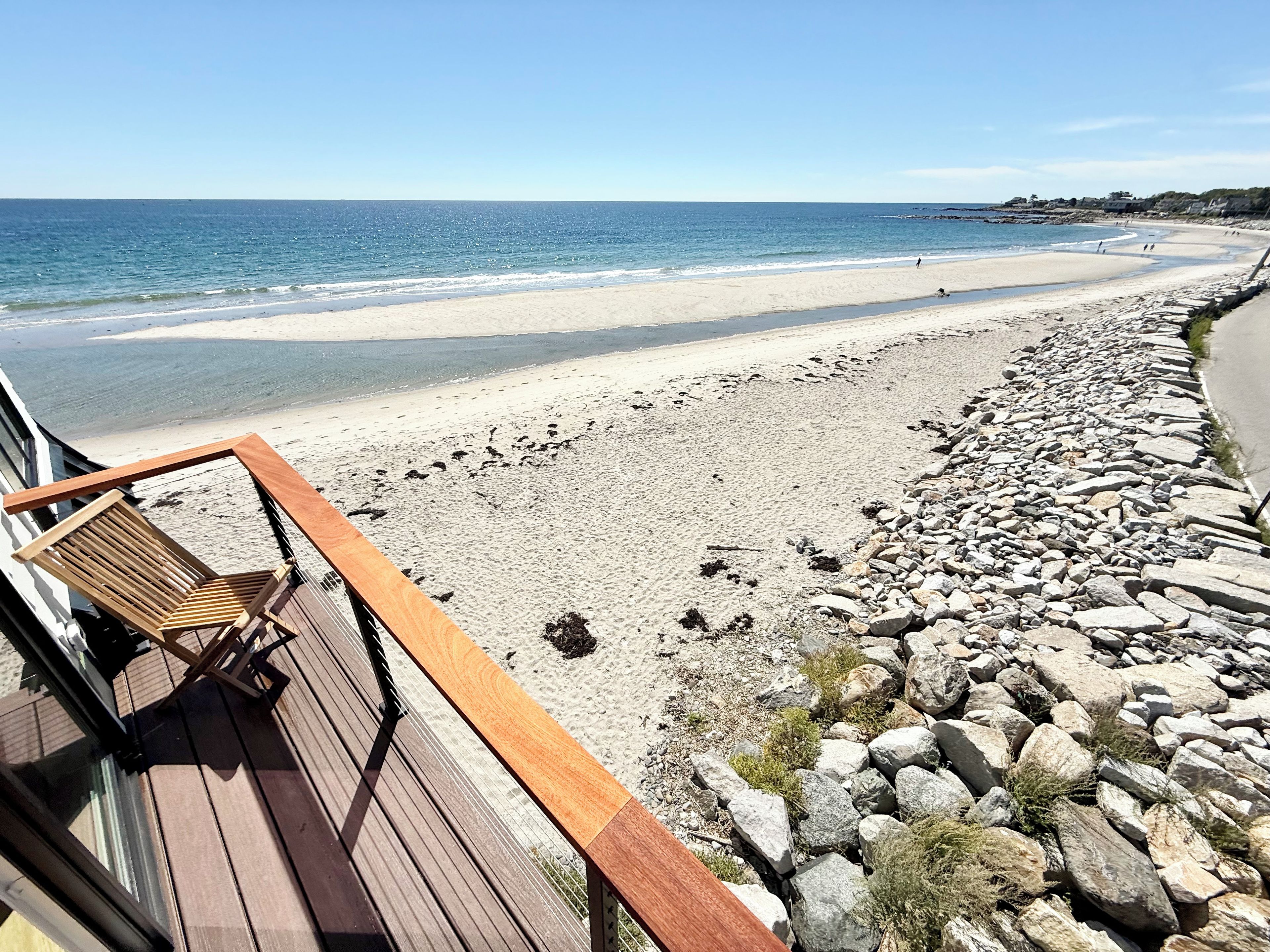 A beach with a wooden deck and a chair, overlooking a sandy beach and rocky shoreline.