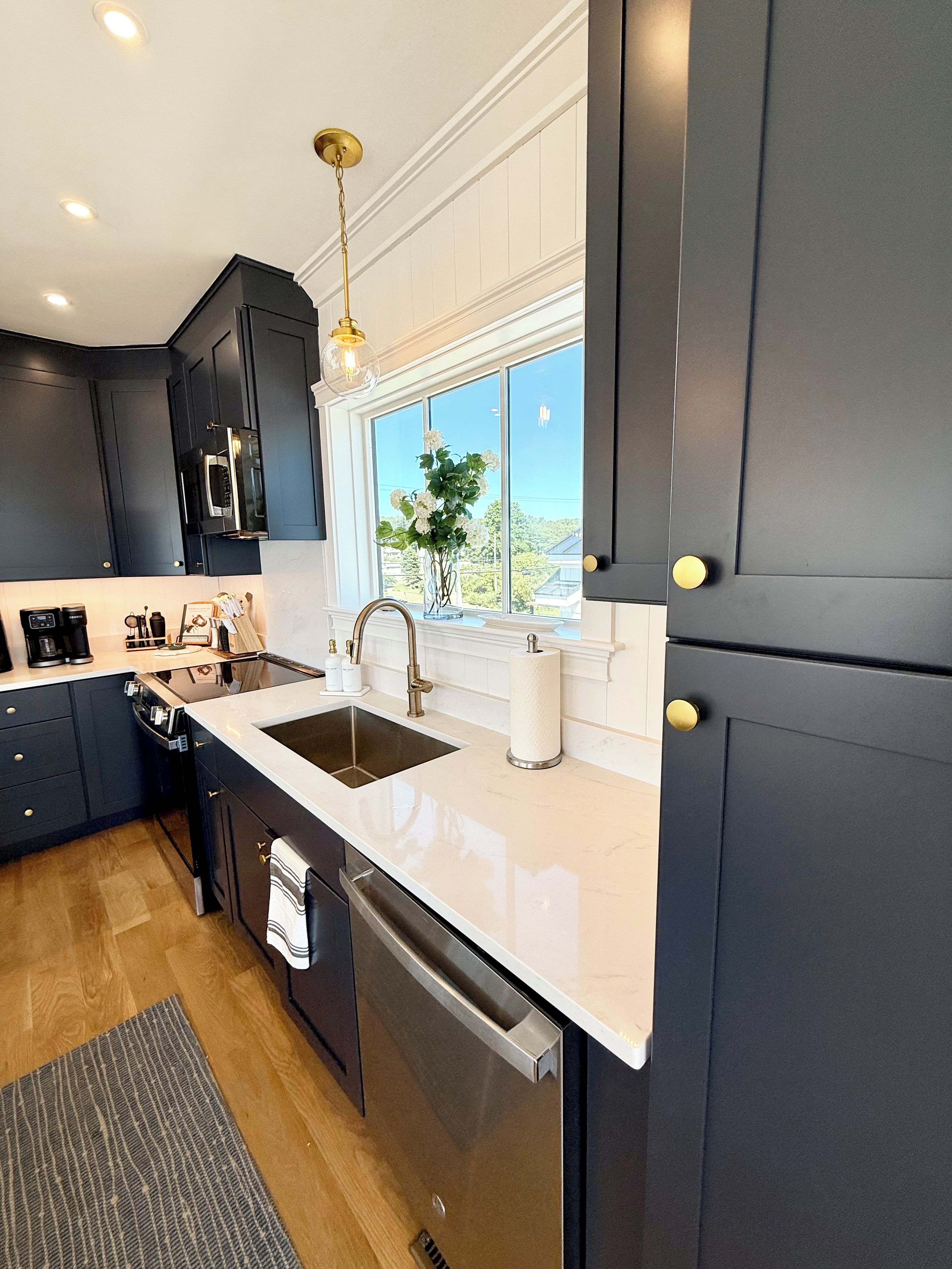A modern kitchen with dark cabinets, a white countertop, stainless steel appliances, and a window with a view.