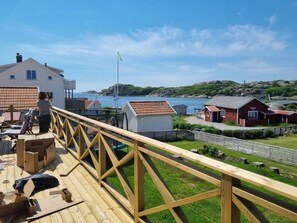 Una terraza de madera con barandilla que da a la costa, un cobertizo pequeño y un edificio con techo rojo.