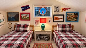 A bedroom with two beds, a window with blinds, a lamp, and various framed pictures on the wall. - Baby Island Bungalow (Langley)