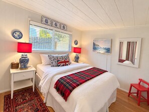 A bedroom with a bed, two bedside tables, a red lamp, a red chair, a window with blinds, and a painting on the wall. - Baby Island Bungalow (Langley)