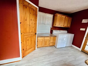Une cuisine de taille réduite, dotée de meubles en bois, d’un lave-linge et d’une fenêtre avec des stores.
