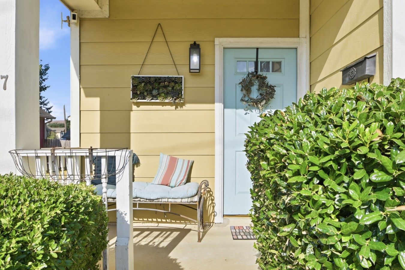 A porch with a cushioned bench, a hanging planter, and a door with a wreath.