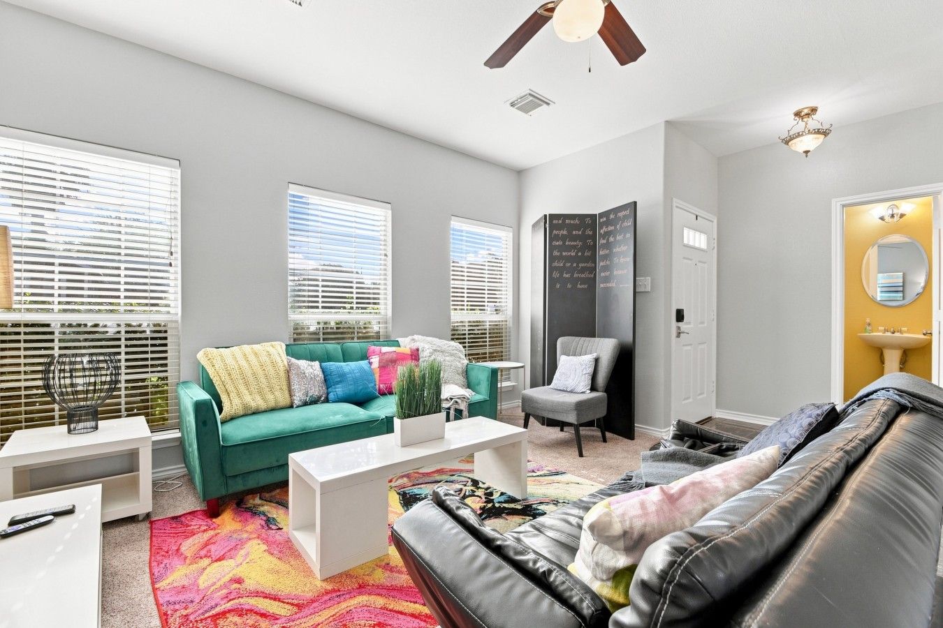 A modern living room with a green sofa, a black leather couch, and a white coffee table. There is a window with blinds, a ceiling fan, and a small white table with a lamp.