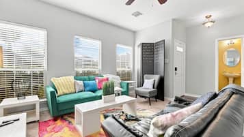 A modern living room with a green sofa, a black leather couch, and a white coffee table. There is a window with blinds, a ceiling fan, and a small white table with a lamp.