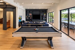 A modern living room with a pool table, a dark entertainment unit, and a large window offering a view of the outdoors. - Villa Solara | Pool, Sauna, Hot Tub & Sport Court (Miami)