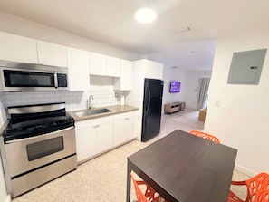 A modern kitchen with stainless steel appliances, a dining area with a wooden table and orange chairs, and a flat-screen TV mounted on the wall. - Stylish 2BR Near Wynwood | Pool + Free Parking (Miami)