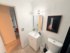 A bathroom with a white sink, mirror, and toilet. There is a framed artwork on the wall and a small plant on the sink. - Stylish 2BR Near Wynwood | Pool + Free Parking (Miami)