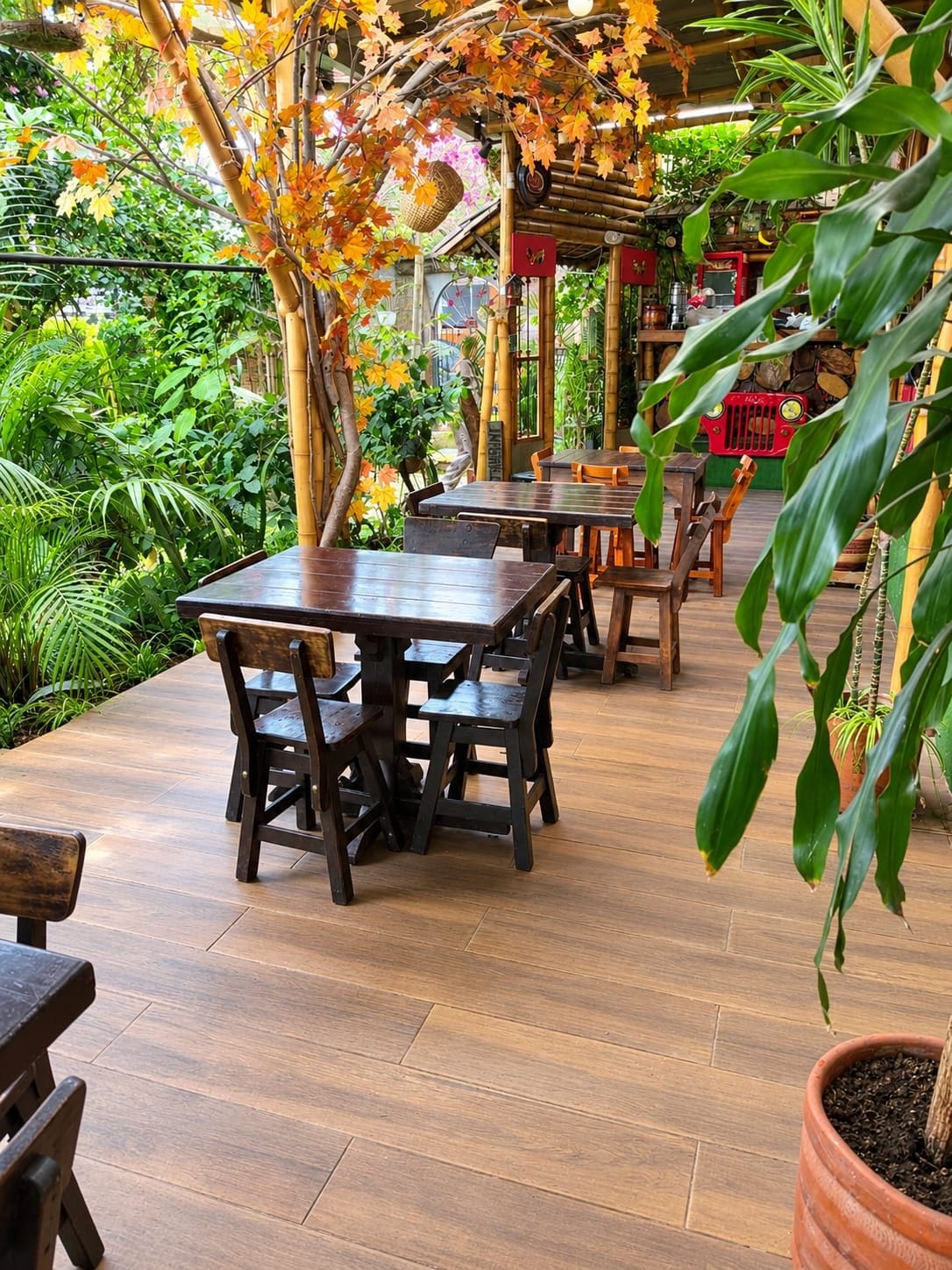 Een terras buiten met houten tafels en stoelen, omringd door weelderige begroeiing en bamboestructuren.