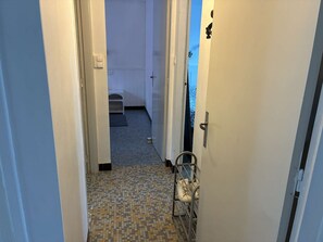 A narrow hallway with a tiled floor, a metal bench, and a door leading to a room with a bed. - Les Carolines de Sion (SAINT HILAIRE DE RIEZ)