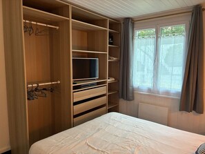 Un dormitorio con cama, un armario de madera con varios estantes, un televisor sobre un mueble bajo y una ventana con cortinas.