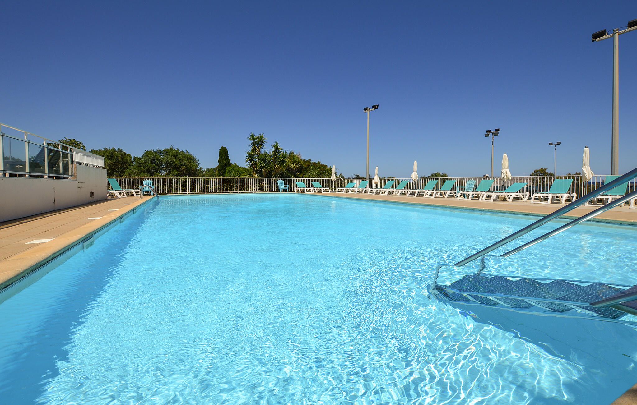 A clear swimming pool with a metal ladder, surrounded by lounge chairs and umbrellas, with a clear sky above.