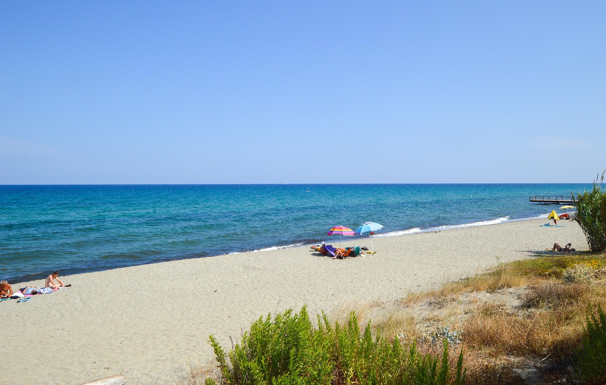 A beach with clear skies, a sandy shore, and a pier extending into the water.