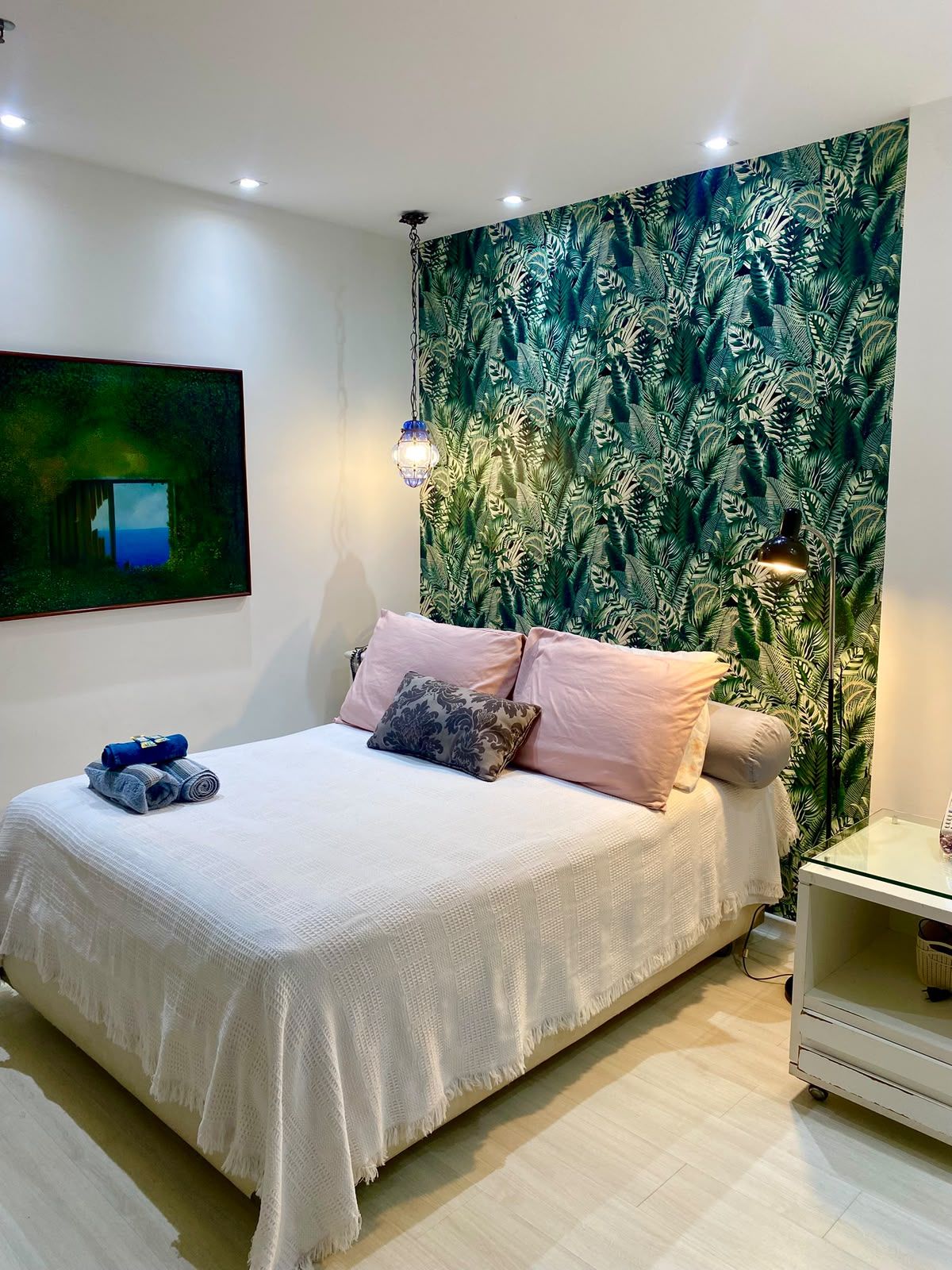 A bedroom with a bed, pillows, a blanket, a wall tapestry, a painting, a lamp, and a bedside table.