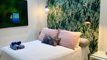 A bedroom with a bed, pillows, a blanket, a wall tapestry, a painting, a lamp, and a bedside table.