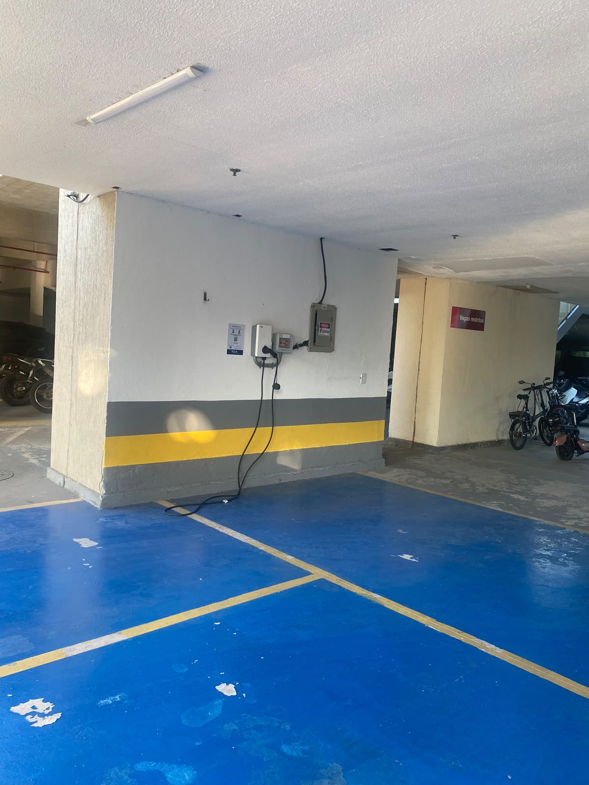 A parking garage with blue painted parking spaces, a wall-mounted electrical box, and a sign reading 'Vagas reservas'.