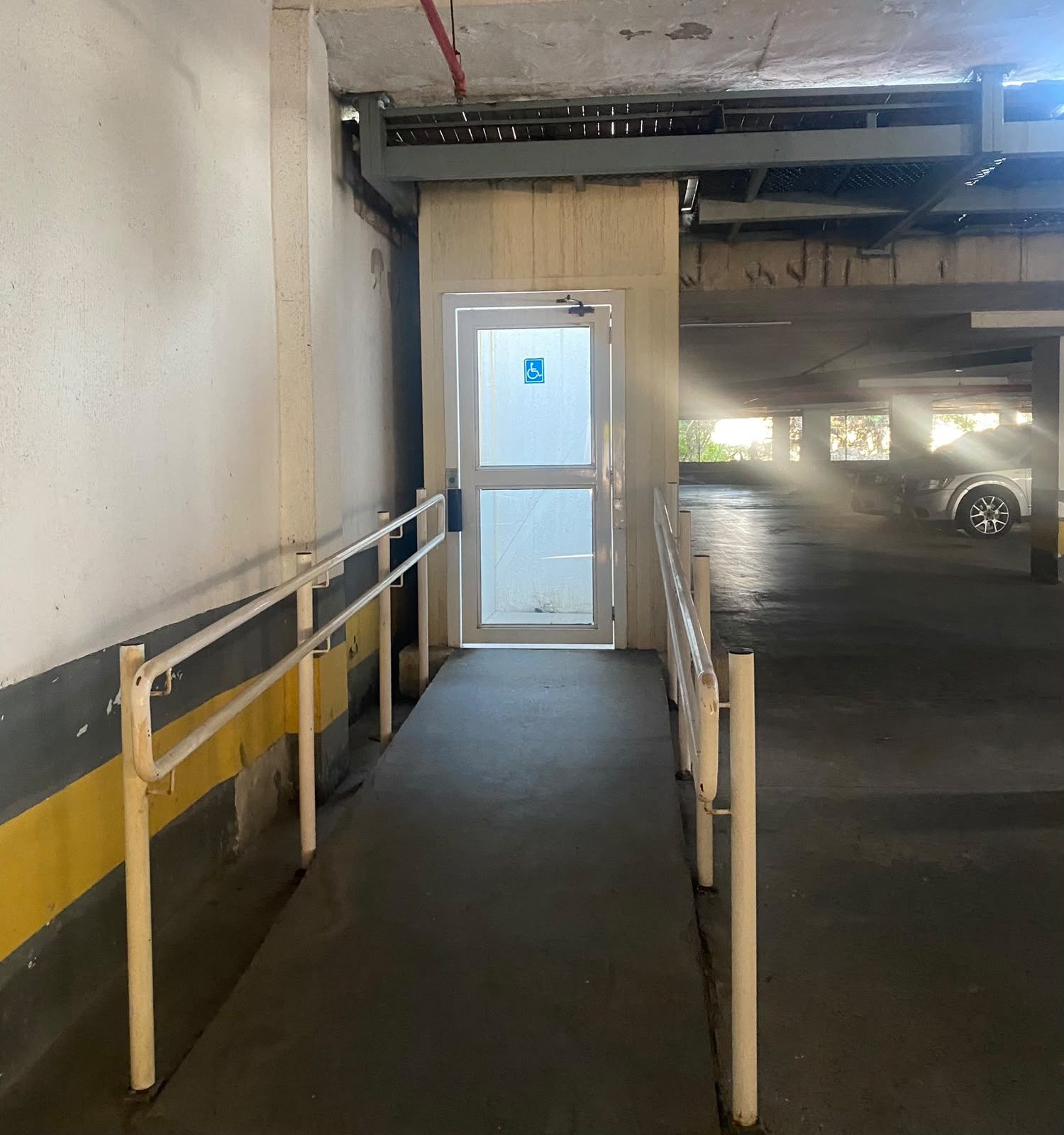 Entrada de um estacionamento com porta acessível para cadeiras de rodas, paredes de concreto e guarda-corpo metálico.
