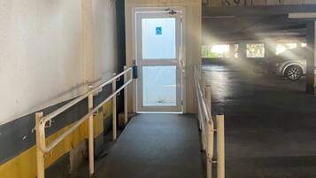 Entrada de um estacionamento com porta acessível para cadeiras de rodas, paredes de concreto e guarda-corpo metálico.