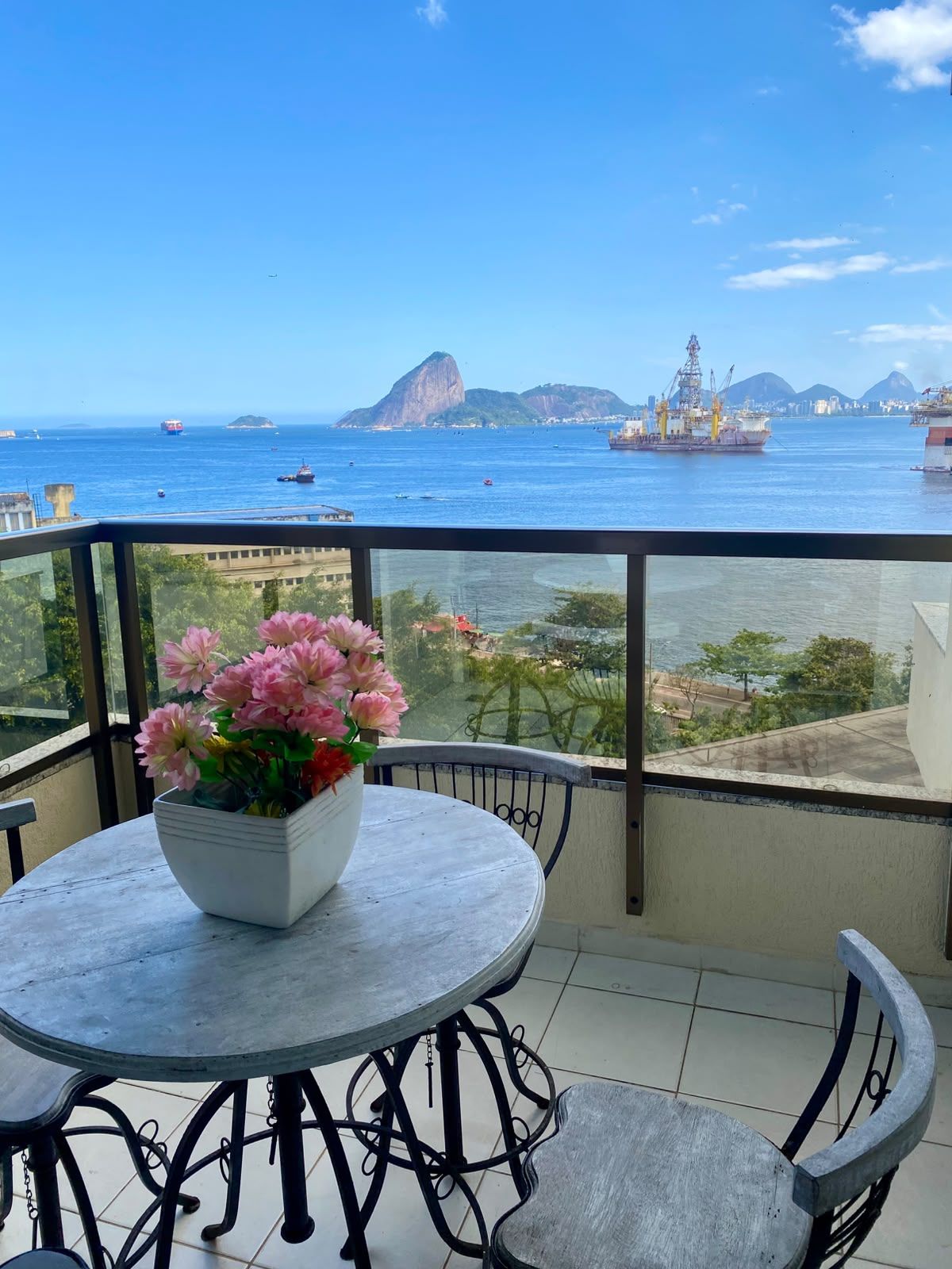 Varanda com mesa e cadeiras, oferecendo vista para a costa, com navios e montanhas.