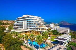 Ein mehrstöckiges Hotel mit Schwimmbereich, umgeben von viel Grün und Blick auf ein Gewässer.