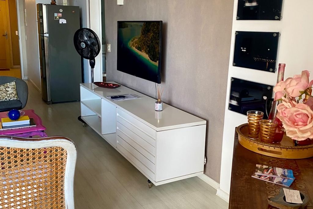 A modern living room with a flat-screen TV mounted on the wall, a white cabinet with drawers, a fan, and a small table with decorative items.
