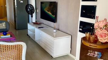 A modern living room with a flat-screen TV mounted on the wall, a white cabinet with drawers, a fan, and a small table with decorative items.