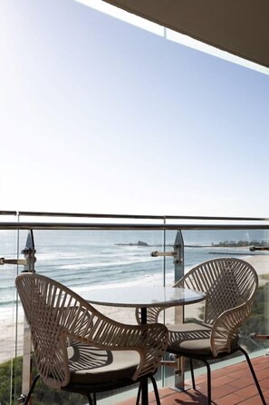 Ein Balkon mit zwei Stühlen und einem Tisch, mit Blick auf einen Strand, das Meer und entferntes Land.