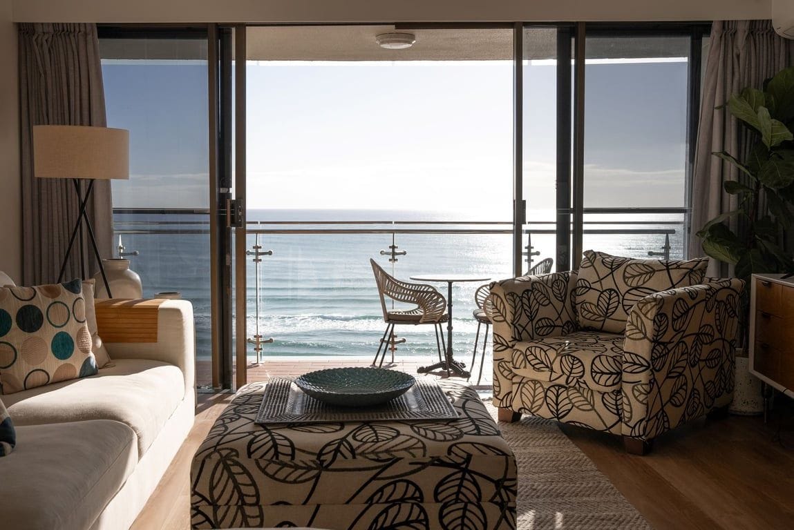 A room with a large sliding glass door leading to a balcony with a view of the sea. There is a sofa, a patterned armchair, and a dining table with chairs.