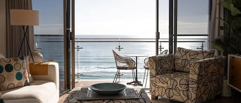 A room with a large sliding glass door leading to a balcony with a view of the sea. There is a sofa, a patterned armchair, and a dining table with chairs.