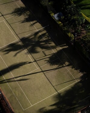 Vista aérea de uma quadra de tênis, com sombras de árvores e uma cerca.