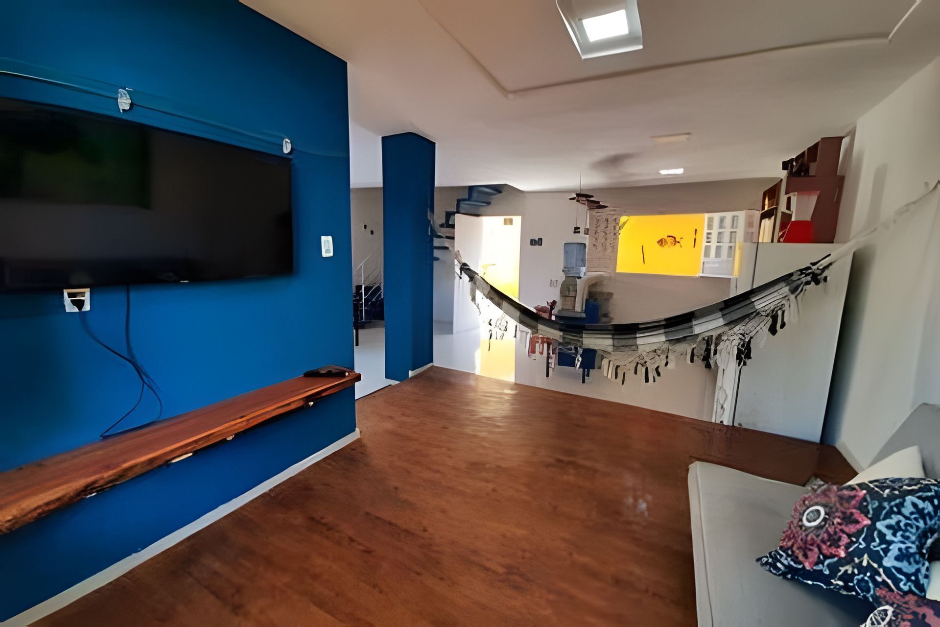 Une pièce avec un plancher en bois, un mur bleu, un téléviseur fixé au mur, un hamac et un canapé agrémenté d’un coussin à motifs.