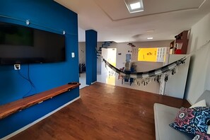 Una habitación con piso de madera, pared azul, televisor montado en la pared, una hamaca y un sofá con un cojín estampado.