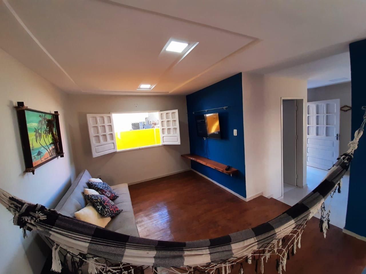 A room with a hammock, a sofa, a TV on the wall, and a window with blinds.