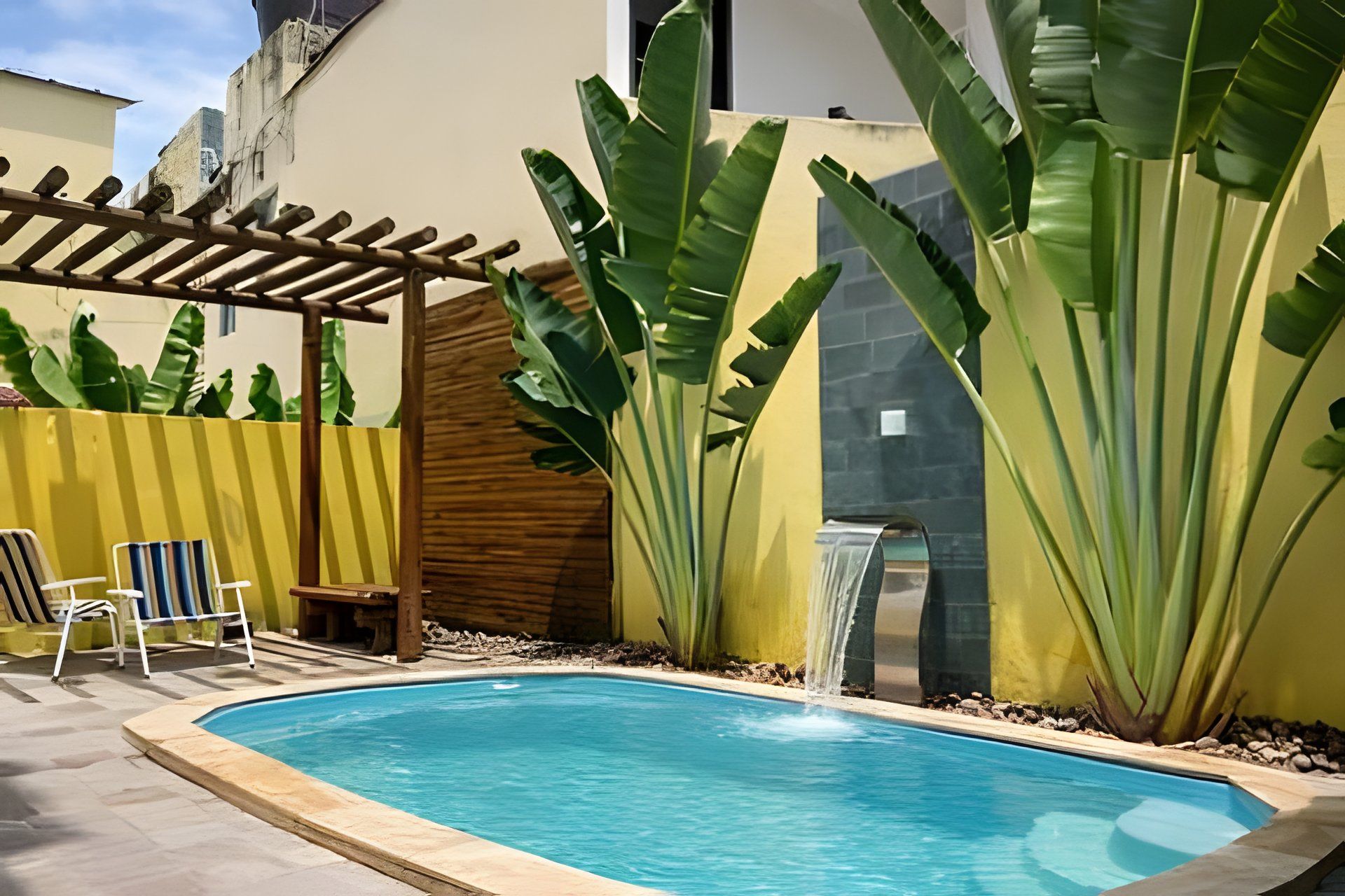 A swimming pool with a wooden pergola, two lounge chairs, and large banana plants.