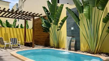 A swimming pool with a wooden pergola, two lounge chairs, and large banana plants.