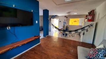 A living room with a flat-screen TV mounted on a blue wall, a wooden bench, a gray sofa with patterned cushions, and a hammock in the background.
