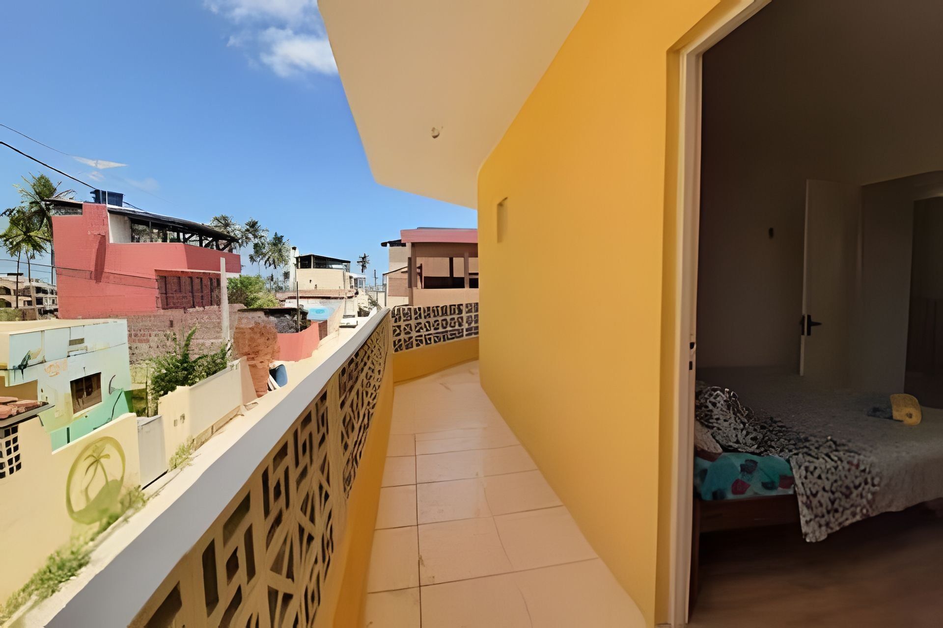 Uma varanda com piso de azulejos, uma cama com colcha estampada e vista para edifícios vizinhos.