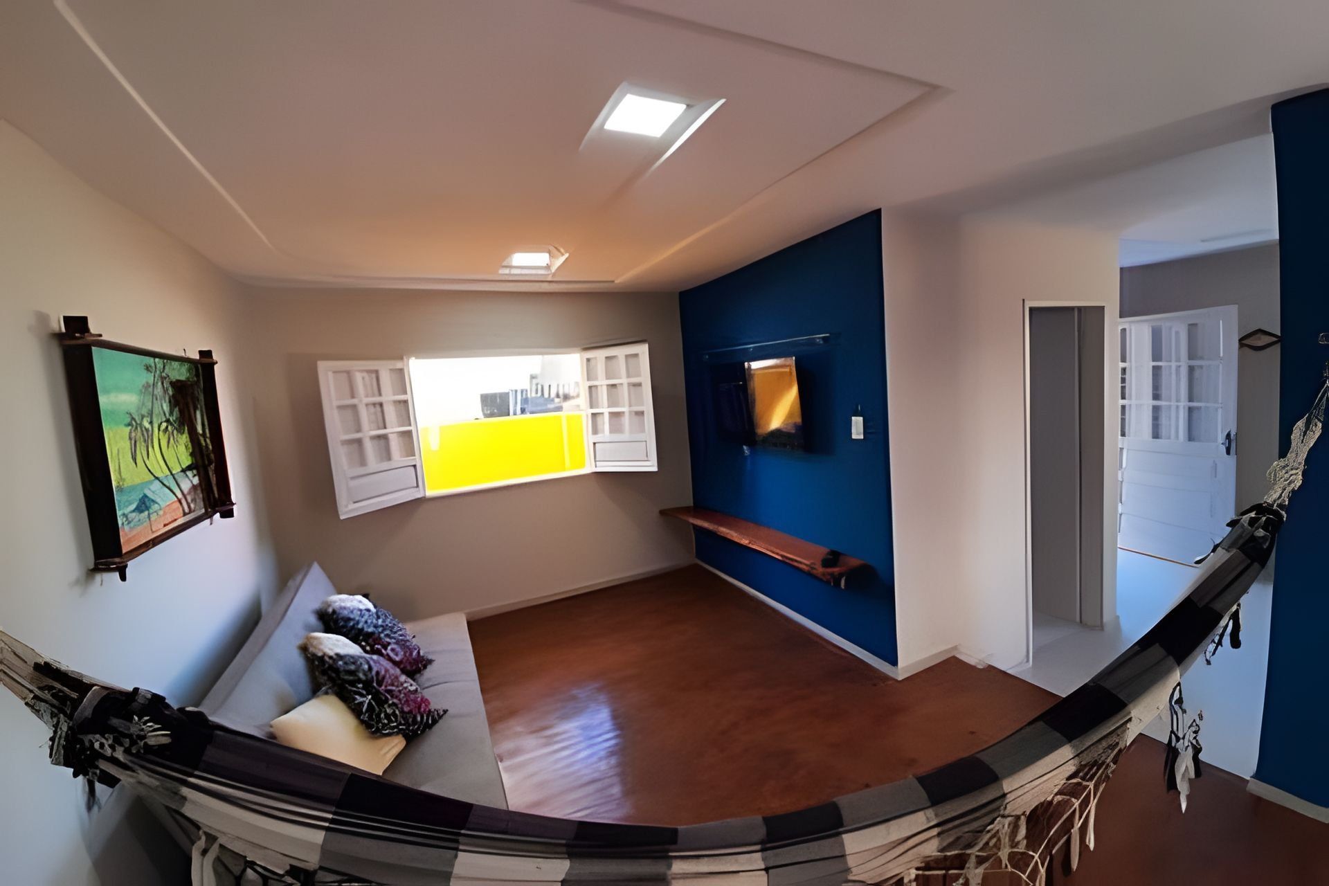 A hammock in a room with a wooden floor, a bench, a wall-mounted TV, and a door leading to another room.