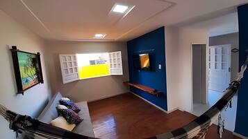 A hammock in a room with a wooden floor, a bench, a wall-mounted TV, and a door leading to another room.